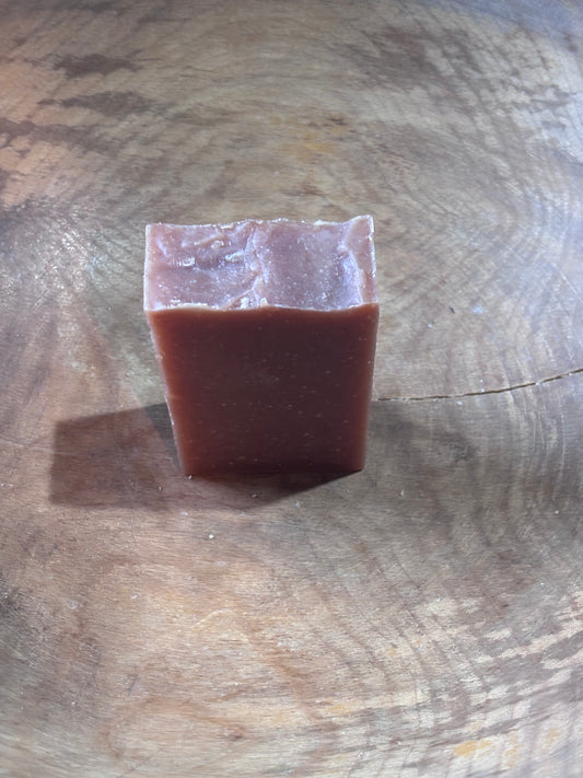A red bar of handmade soap in a wooden bowl. Cherry Almond Scented. Ingredients include distilled water, olive oil, palm oil, coconut oil, castor oil, sodium hydroxide (lye), fragrance oil and madder root powder.
